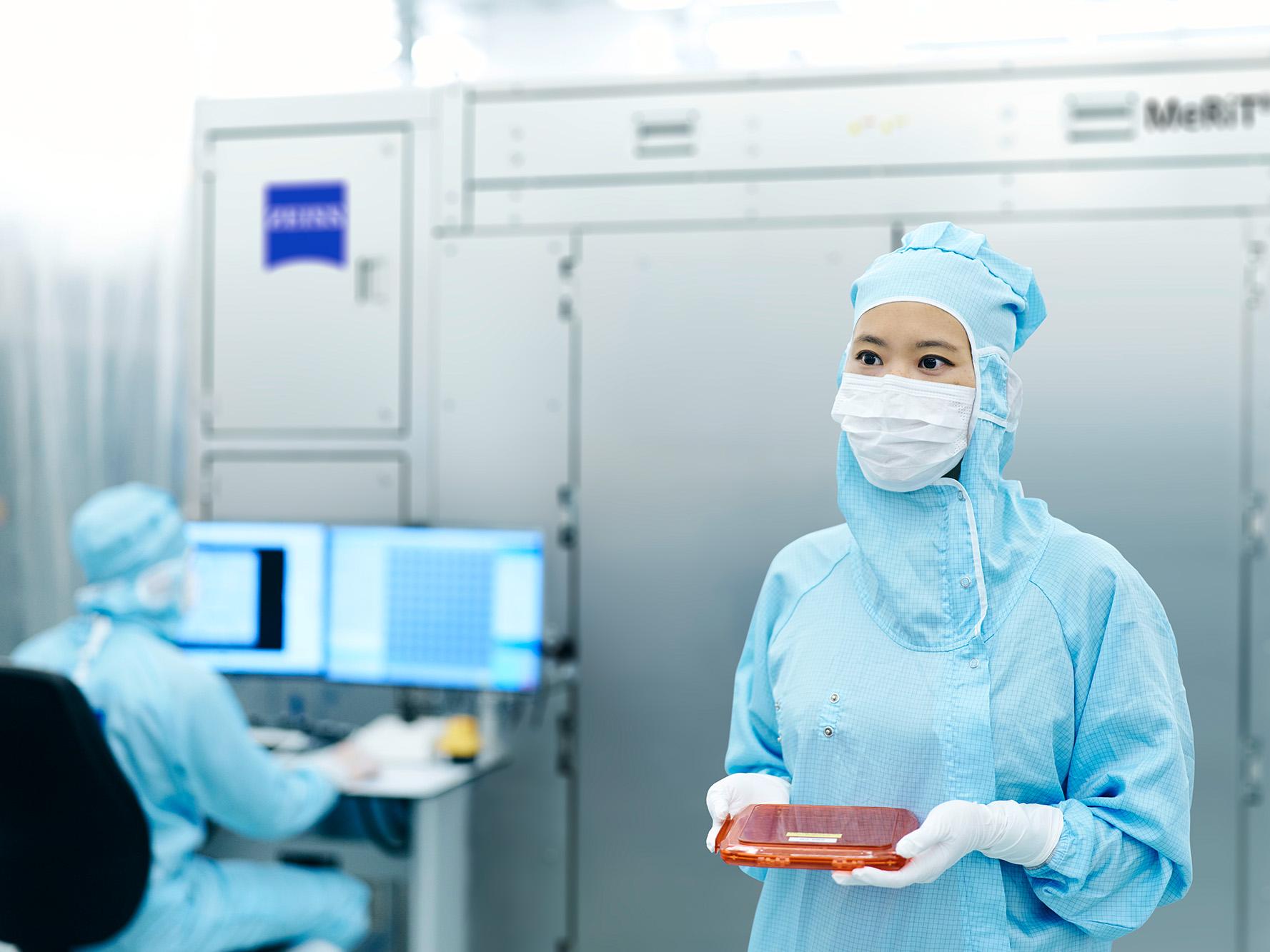 一名員工在潔淨室中手持光罩，其同事則在電腦前工作。