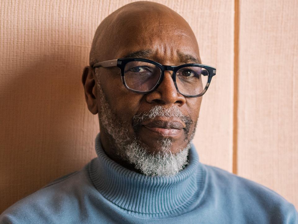 Older man with black glasses and a gray beard looks skeptical.He is wearing a blue turtleneck sweater